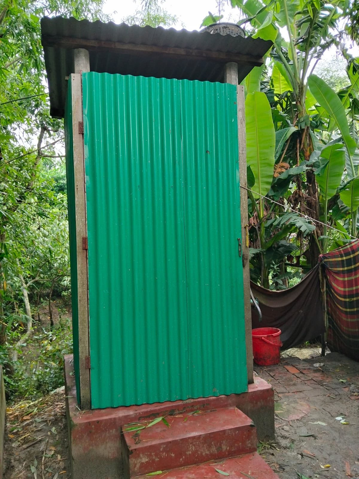 Green tin latrine on a raised base with brick steps, banana plants behind.